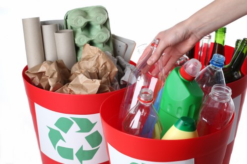 A pair of bright red recycling bins with white labels featuring the green recycling symbol, placed outdoors on a paved surface. The left bin contains several brown paper bags, crumpled and filled with cardboard and paper waste, along with a set of light green plastic egg cartons. The right bin is filled with clear plastic bottles, some with colored caps in red, green, or blue, and a few plastic containers, including a yellow and green detergent bottle. A person's hand, with fingers partially visible and wearing a short-sleeved shirt, is seen reaching into the right bin, indicating active sorting or placing of waste materials. In the background, a section of a driveway or paved area is visible, suggesting a residential or garden clearance setting, possibly in New Cross. The scene exemplifies typical rubbish separation and collection activities related to garden or household waste removal services by Garden Clearance New Cross.