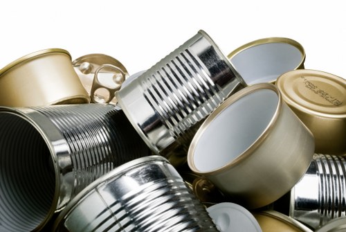 A close-up image showing a pile of discarded tin cans and aluminum containers, some with smooth metallic surfaces in silver, gold, and light grey tones, scattered across a flat surface. The cans include both open and closed lids, with some stacked or leaning against each other, displaying ribbed textures on the sides and shiny reflective finishes. The background is neutral and out of focus, emphasizing the assortment of metallic rubbish, which aligns with waste collection and rubbish removal services. This visual is relevant to property or garden clearance in New Cross, where professional rubbish removal companies like Garden Clearance New Cross handle the disposal of such waste materials efficiently.