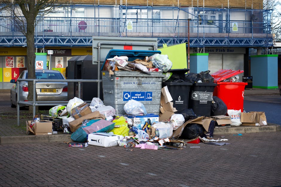 A sanitation worker in a yellow and orange safety vest is operating a red rubbish collection vehicle on a paved roadside area, with the back of the vehicle open for waste loading. The worker appears t